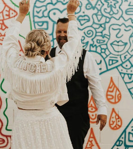 Bride wearing bridal jacket with fringe that says hitched on the back.